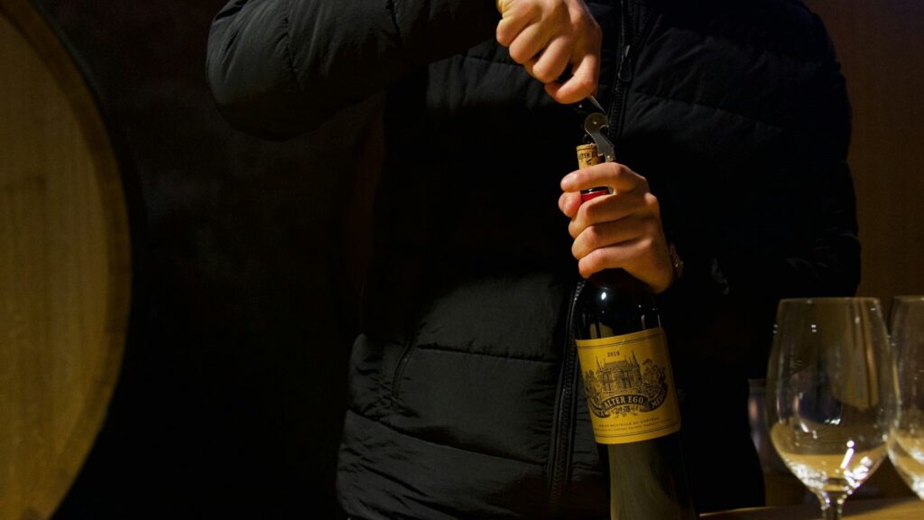 Person opening a bottle of red wine with a corkscrew beside wine glasses in a softly lit cellar, illustrating the question does wine expire?